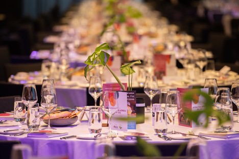 Image of round table setting from NAWIC International Women’s Day with plant in the middle of table, drink glasses and empty table settings before guests arrive. Background of image is a blurred representation of the tables that are behind them. 