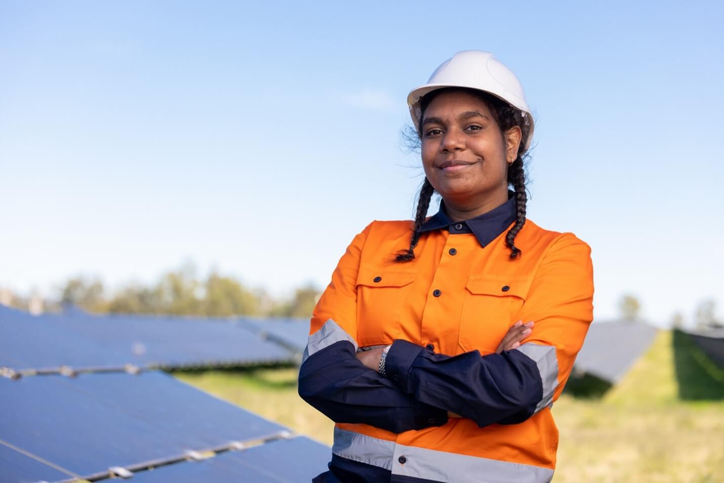 Young Aboriginal female in hard hat and orange hi-vis, standing confidently with arms crossed and smiling at the camera amongst a background of fixed to ground solar panels.